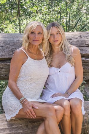 Two people wearing white summer dresses sit together on a wooden bench in a sunny outdoor setting.