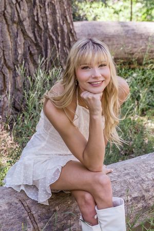 A woman in a white dress sits on a log in a natural outdoor setting with bright sunlight filtering through trees.