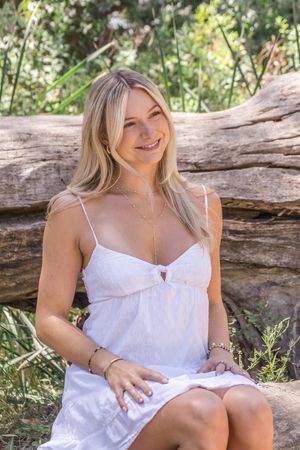 Woman in white summer dress poses on rustic log in dappled sunlight surrounded by green foliage.