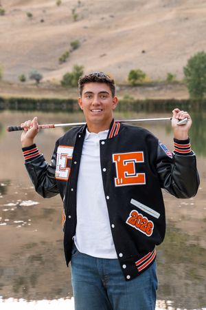 A high school student wearing a varsity letterman jacket with orange 'E' emblem stands near water.