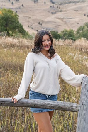 A candid outdoor photoshoot showing someone in a white sweater and denim shorts leaning on a rustic wooden fence.