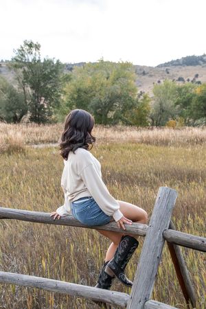 A person in a white sweater and jeans sits on a rustic wooden fence overlooking a golden autumn meadow.