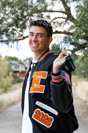 A person wearing a black varsity letterman jacket with orange letters and an American flag patch poses outdoors.