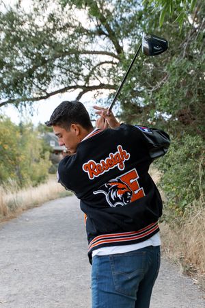 A person wearing a black varsity jacket with orange accents stands on a gravel path by overgrown brush and trees.