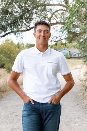 A person in a white polo shirt and blue jeans poses confidently outdoors on a dirt path with trees in the background.