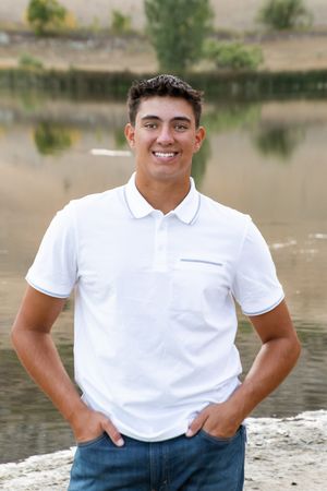 Person in white polo shirt and jeans posed near a lake with trees in the background.