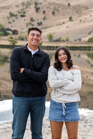 A couple poses playfully by a lake with barren hills in the background wearing casual black and white outfits.