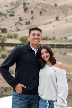 A couple poses together near a lake with hills in the background wearing matching white and dark clothing.