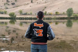 Letterman jacket with orange E patch seen from behind at a serene lakeside setting.