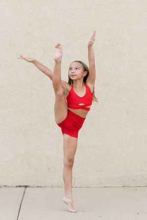 Young gymnast in red outfit performing a standing split with arms raised against a beige wall.