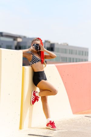 Young woman in polka dot bikini top and high-waisted shorts holds vintage camera, wearing red Adidas sneakers outdoors.