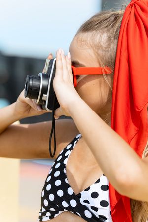 Young blonde girl in polka dot swimsuit and red sunglasses taking photo with vintage film camera outdoors.