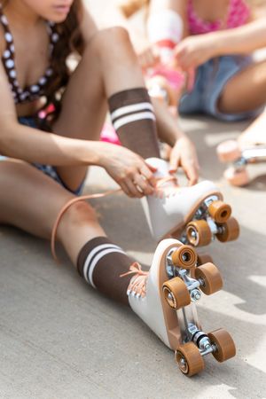 Girl sitting on ground lacing up white and brown retro quad roller skates while wearing brown knee-high socks.