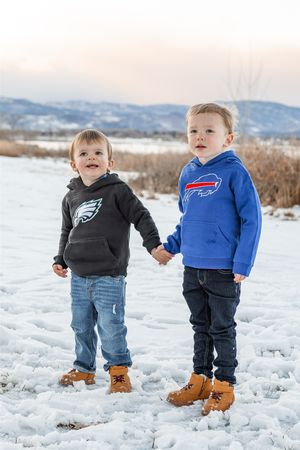 Two children wearing sports team hoodies stand together holding hands in the snow with mountains in the background.