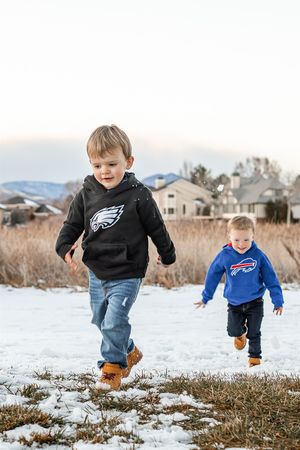 People wearing sports team apparel play and jump in the snow with mountains and houses in the background.