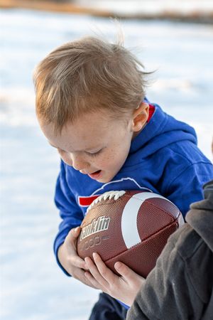 Young child in blue hoodie playfully bites a football while playing in the snow.