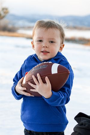 A young child in a blue hoodie holds a football while playing in the snow.