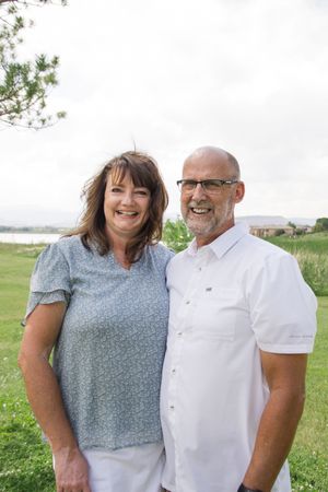 A couple in casual white and blue attire smiles together on a grassy hillside.