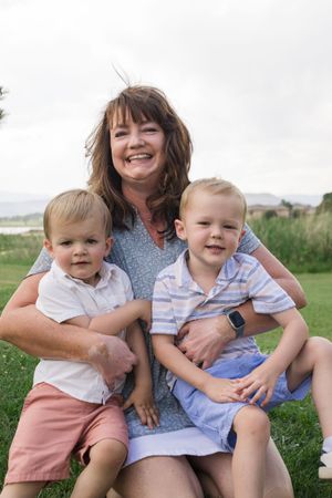 A woman in blue floral top poses with two young children on her lap outdoors.