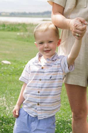 Young child in striped shirt smiles while holding an adult's hand outdoors.