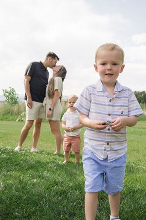 A young child in striped shirt stands in foreground while family poses in background on grassy field.