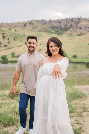 A newly engaged couple posing together in a grassy field with rolling hills.