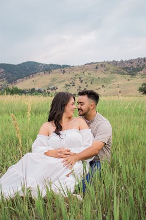 Romantic couple poses in tall grass field with mountains in background during engagement photoshoot.