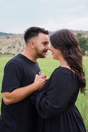 Intimate couple in matching black outfits share a tender moment in a grassy field with rolling hills.