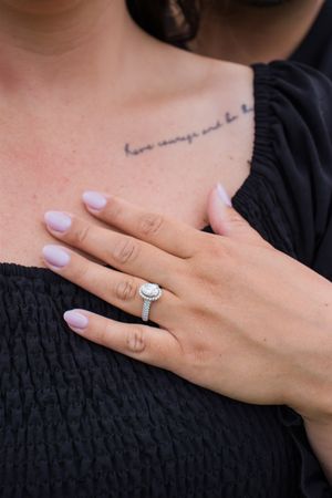 Close-up of engagement ring and script tattoo on woman's collarbone with black fabric texture.