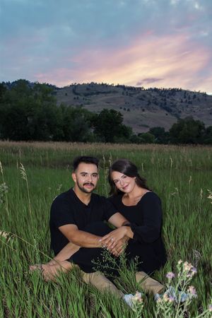 Couple sits in tall grass during sunset with mountains and pink sky in background.