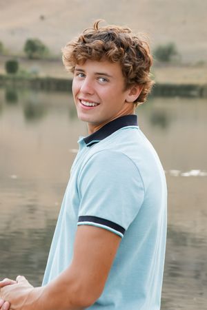 Person in light blue polo shirt poses casually by a serene lake with hills in the background.
