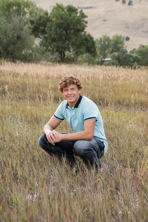 A young person in a light blue polo shirt sits casually in a field of tall grass and wildflowers.