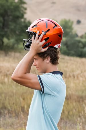 Someone puts on an orange sports helmet while standing in a meadow on a sunny day.