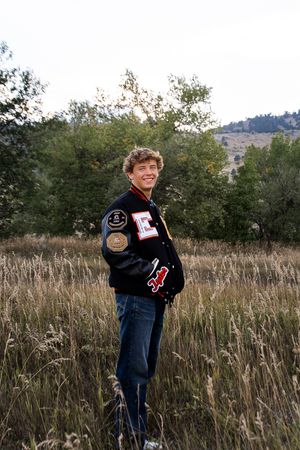 High school student poses in tall grasses wearing a varsity letter jacket at sunset.