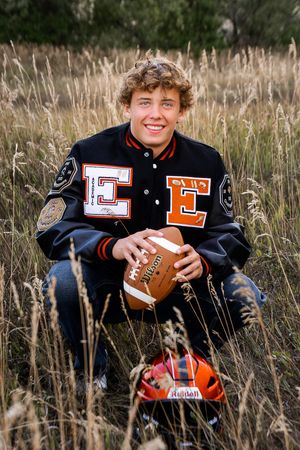 High school football letterman jacket portrait in tall grass with football and helmet, against natural autumn background.