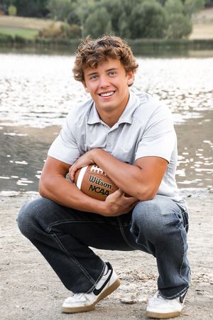 Student athlete crouches by a lake holding a football for senior portraits.