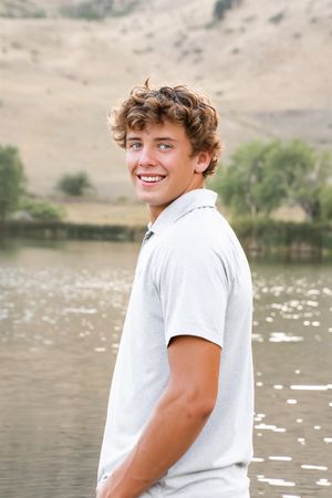 Portrait in white polo shirt smiling by a scenic lake with mountains in background.
