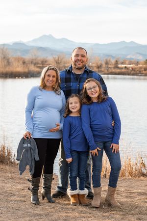 A family wearing coordinated blue outfits poses together by a lake with mountains in the background during autumn.