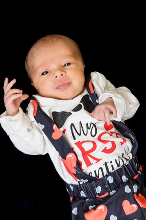 A baby wearing a black and red Valentine's Day themed outfit with hearts lies against a dark background.