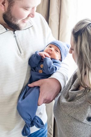 A newborn baby wearing a blue knotted hat and onesie is held lovingly while resting against a beige background.