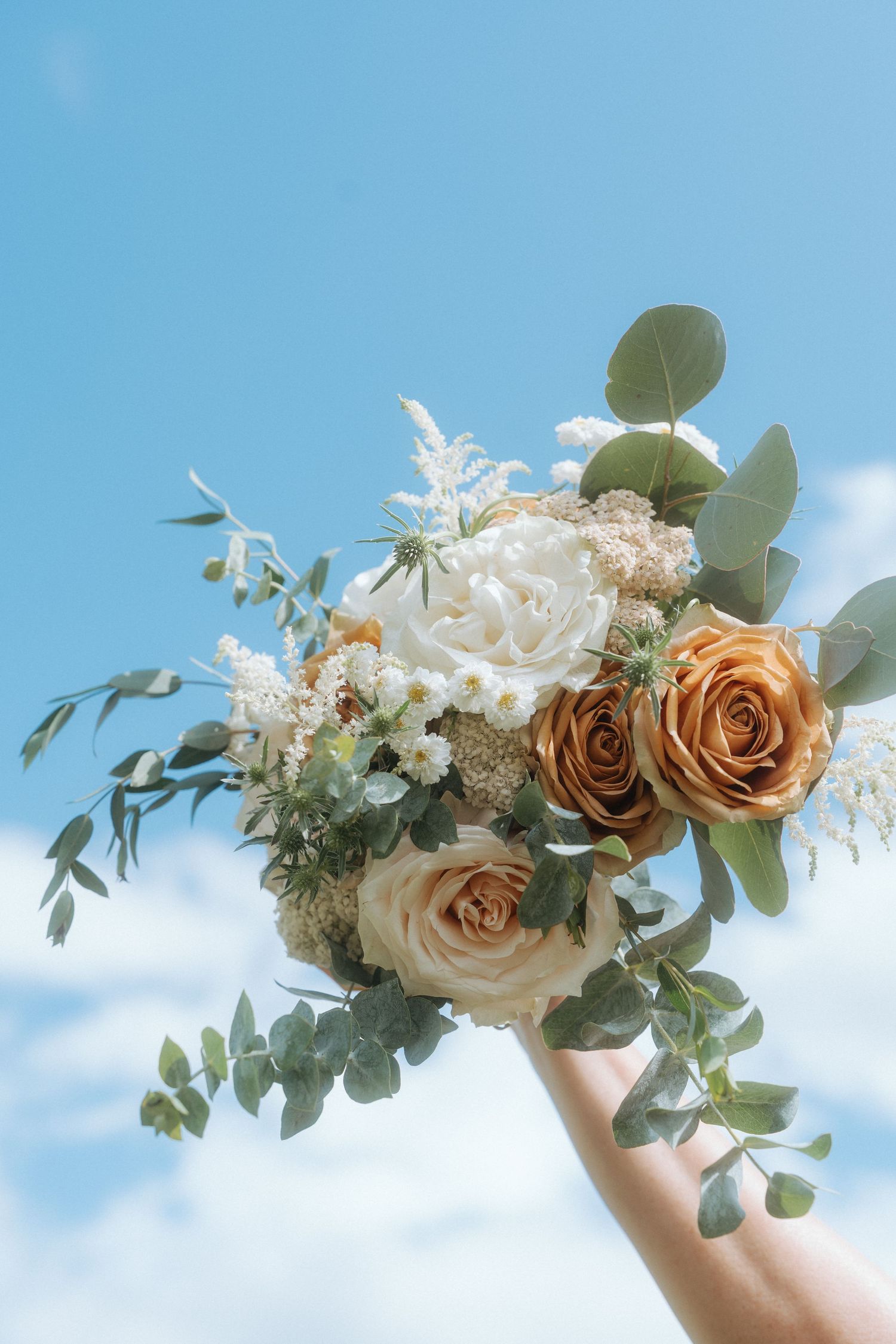 A delicate floral arrangement with peach roses and white flowers held up against a bright blue sky with wispy clouds.