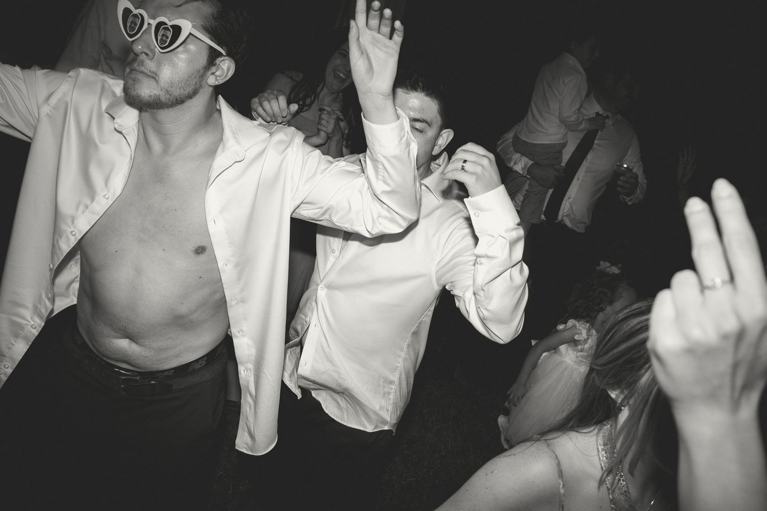 Black and white photo of people dancing energetically at a party with raised arms and open shirts.