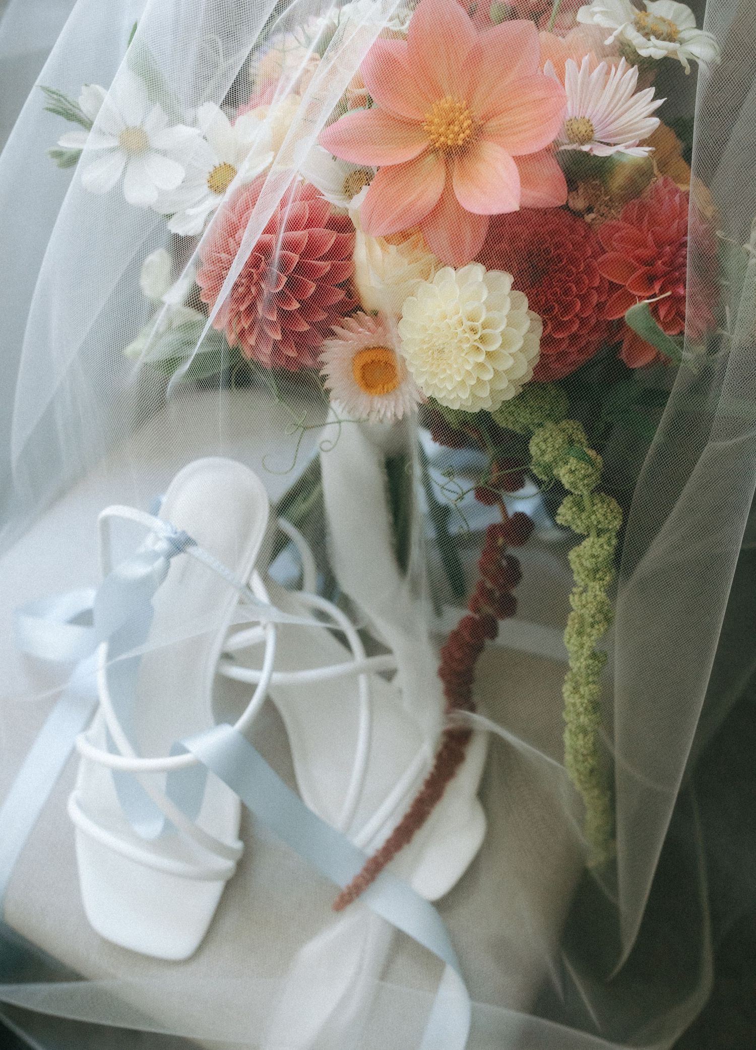 A bridal bouquet of pink, red and white flowers rests beside white sandals through delicate tulle fabric.