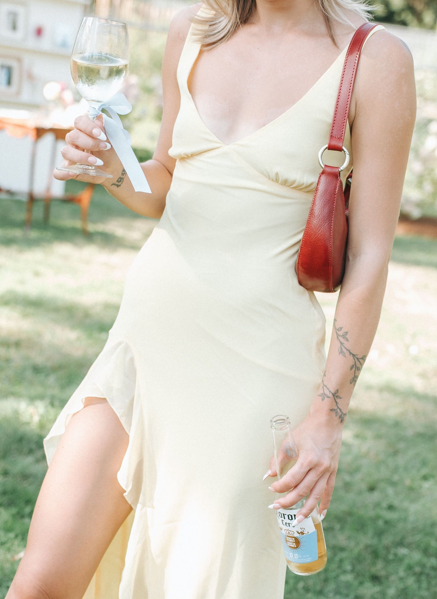 Person in cream-colored slip dress holding wine glass and small bag outdoors on sunny day.