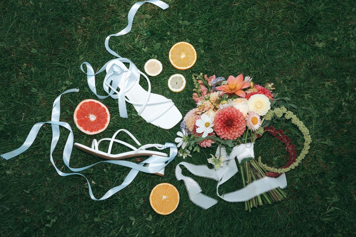Wedding accessories including citrus fruits, white ribbons, and pink floral bouquet arranged on green grass.
