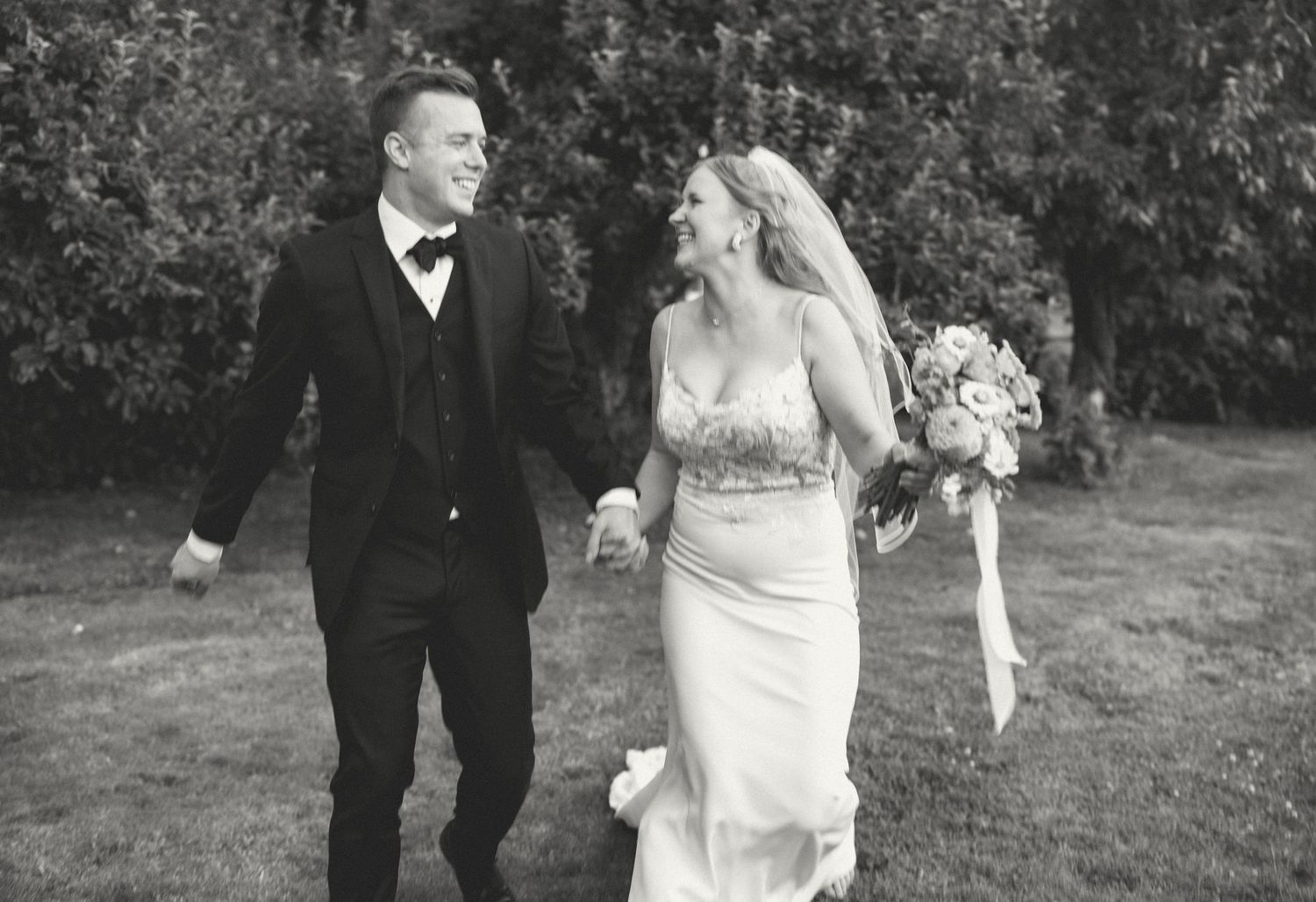 Black and white photo of a couple laughing together while walking outdoors in wedding attire.