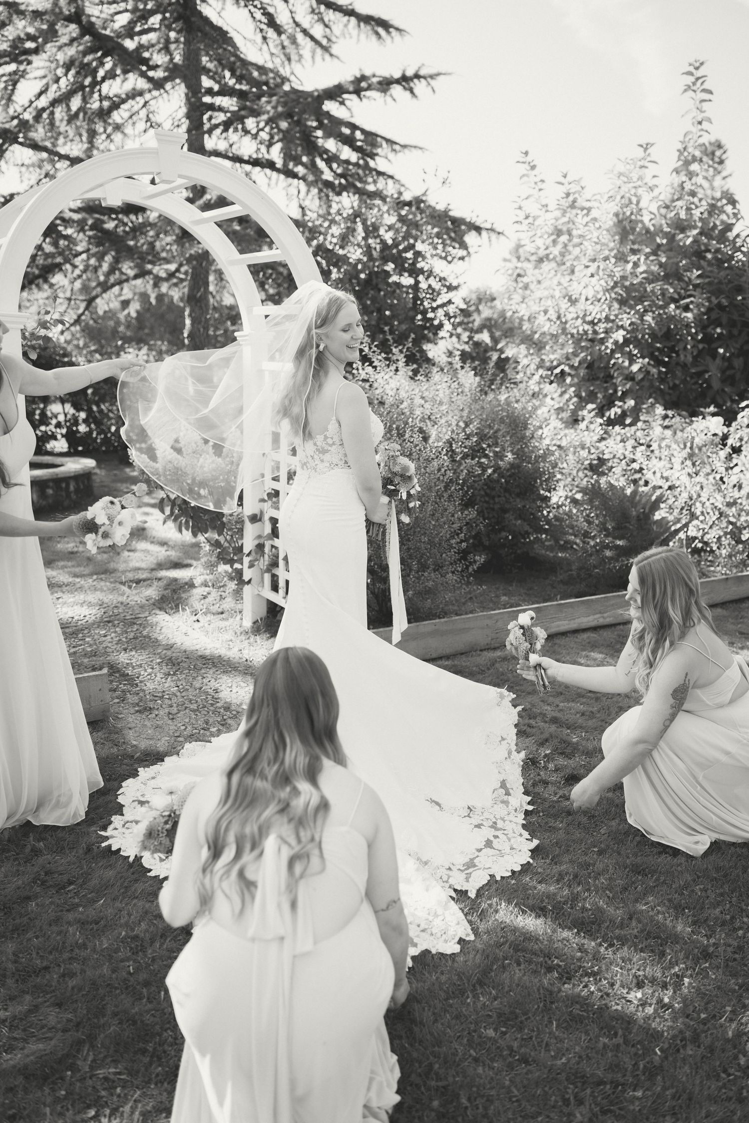 A black and white wedding photo showing a bride in a flowing dress with attendants under an outdoor garden arch.