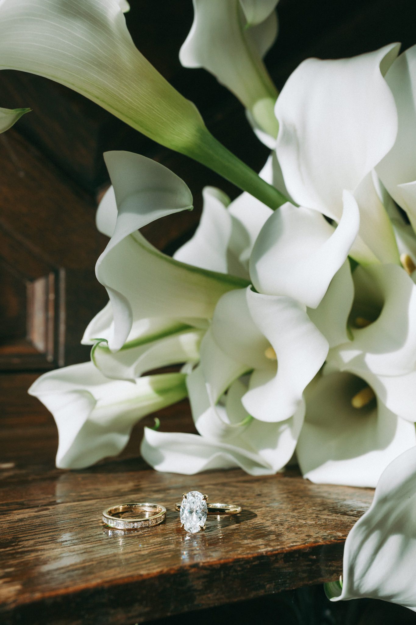 Wedding rings displayed with elegant white calla lilies on wooden surface.