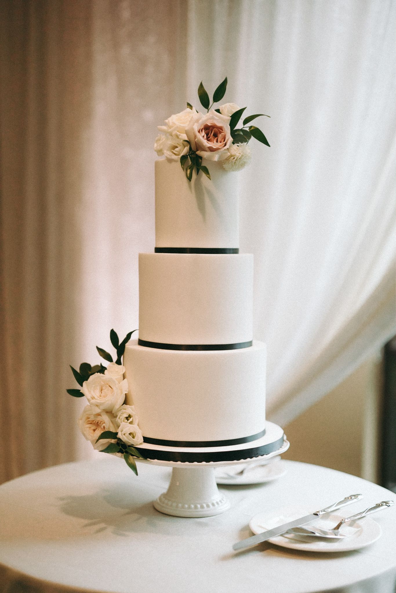 Elegant white wedding cake with black ribbon trim and blush floral accents displayed on a classic cake stand.