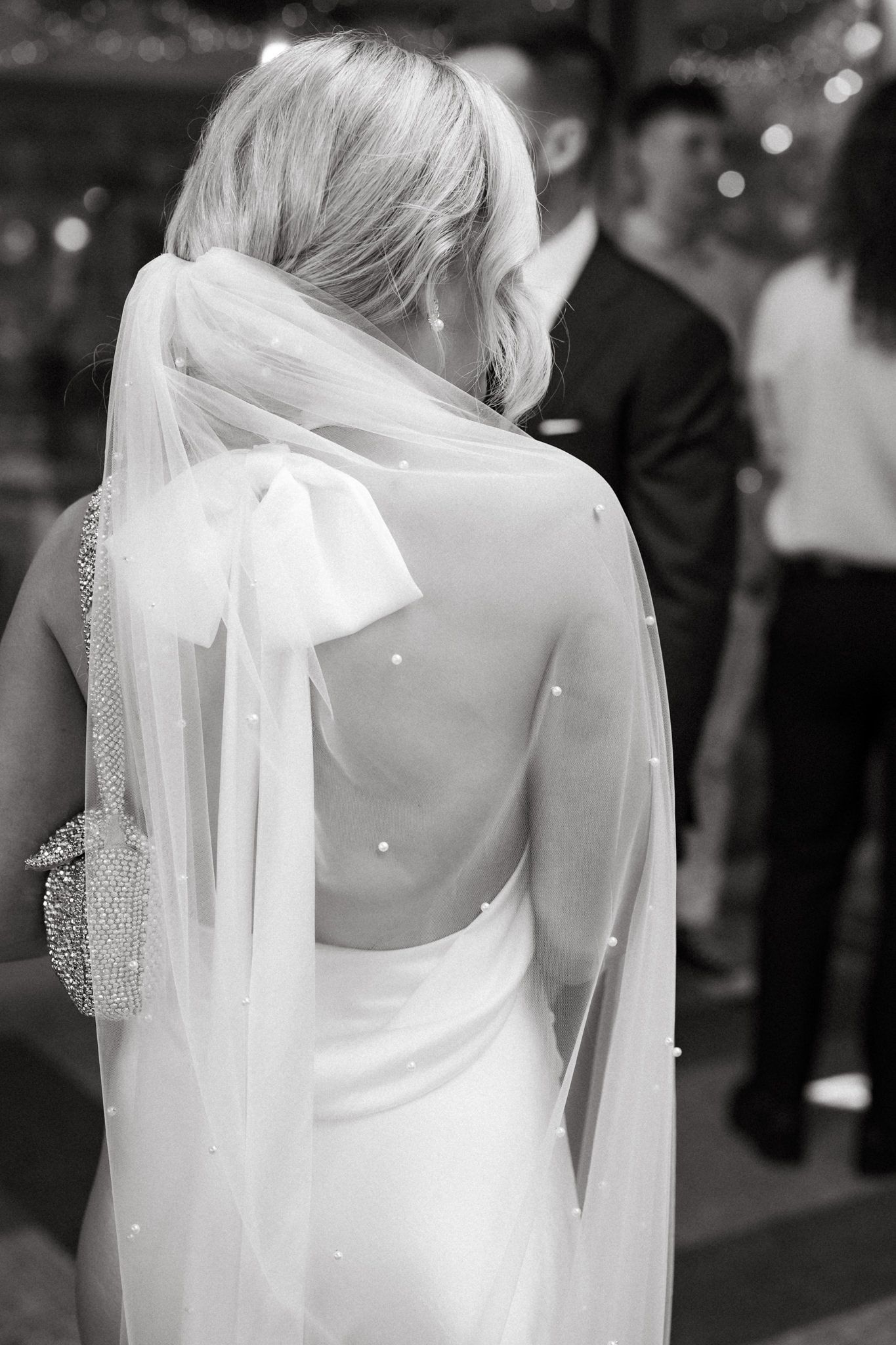 Back view of a bride in a white dress with flowing veil and beaded shoulder details.